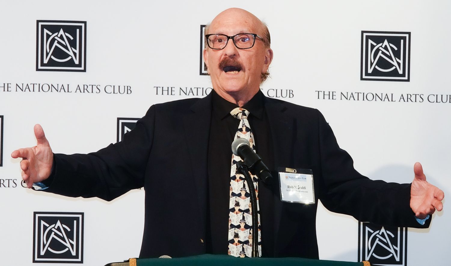 Man with glasses gestures expansively at a podium, National Arts Club backdrop.
