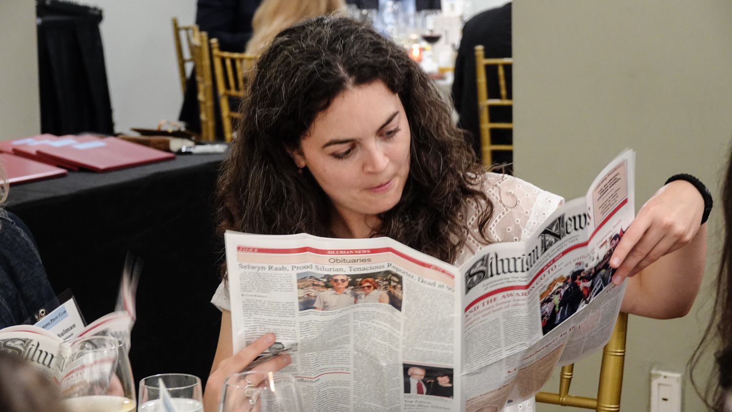 Woman at a table reading a newspaper; indoors, natural light.