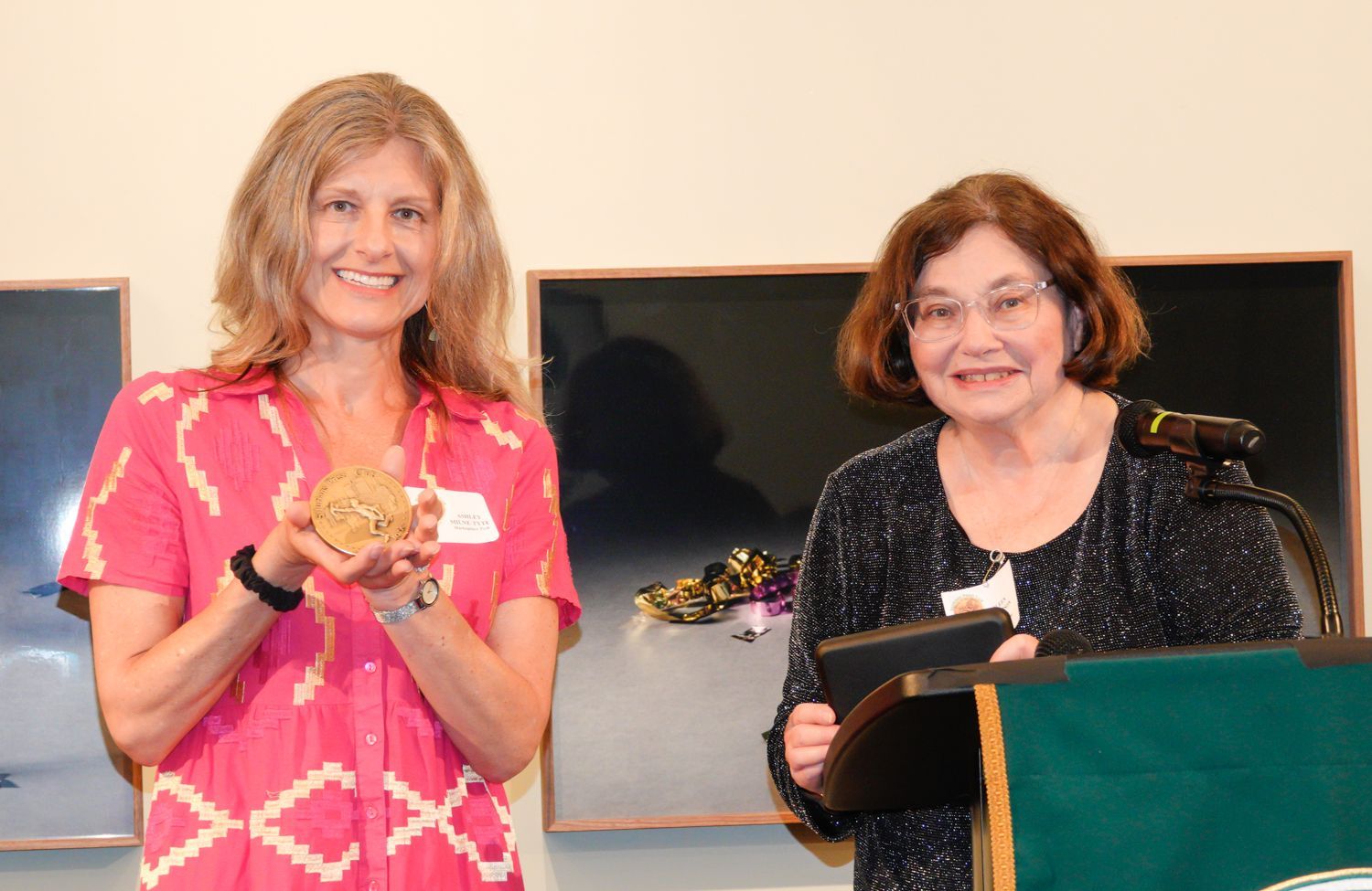Woman holding a medal smiles, another woman speaks at a podium. Two paintings on the wall.