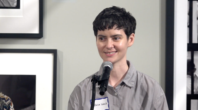 Person with short dark hair smiling in a blue shirt against a gray textured wall