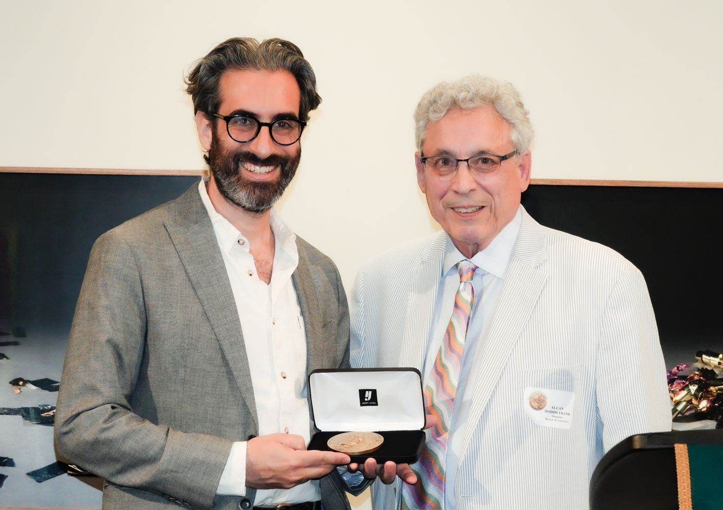 Two men at a presentation: one holding an open gift box with a medal, smiles. The other man smiles, too.