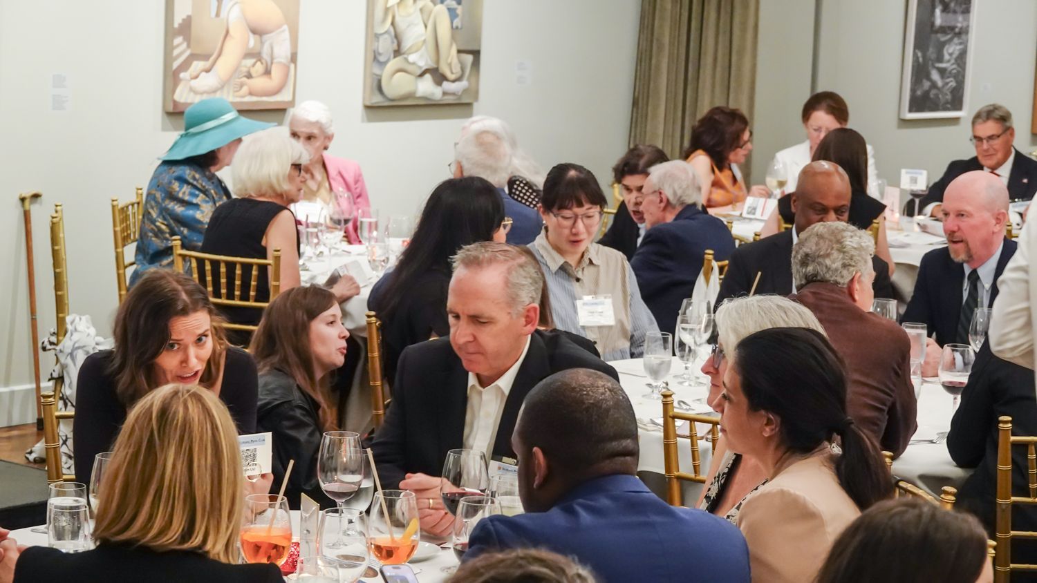 People seated at tables in a room, dining and talking. Artwork hangs on the wall.