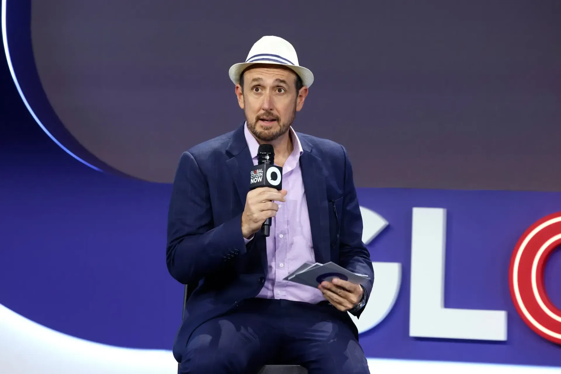 Man speaking on stage into a microphone, wearing a white hat and dark blazer, with a purple event backdrop