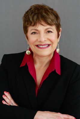Woman with short brown hair, wearing a black blazer over a red shirt, smiling with arms crossed.