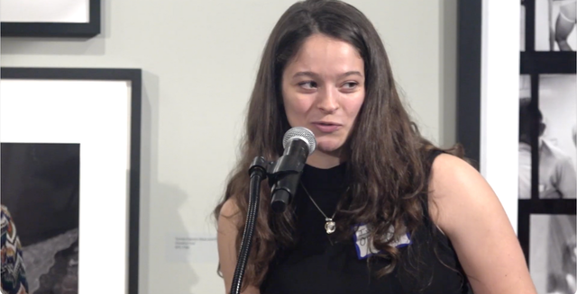 Young person with long brown hair wearing a black turtleneck, standing against a plain light wall.