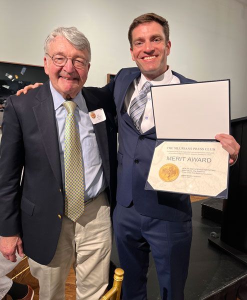 Two men in suits, one holding an award certificate. They're smiling, indoors, possibly at an event.