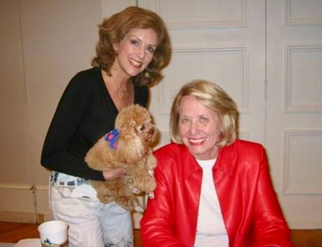 Woman in red jacket smiles with woman holding dog; indoor setting.