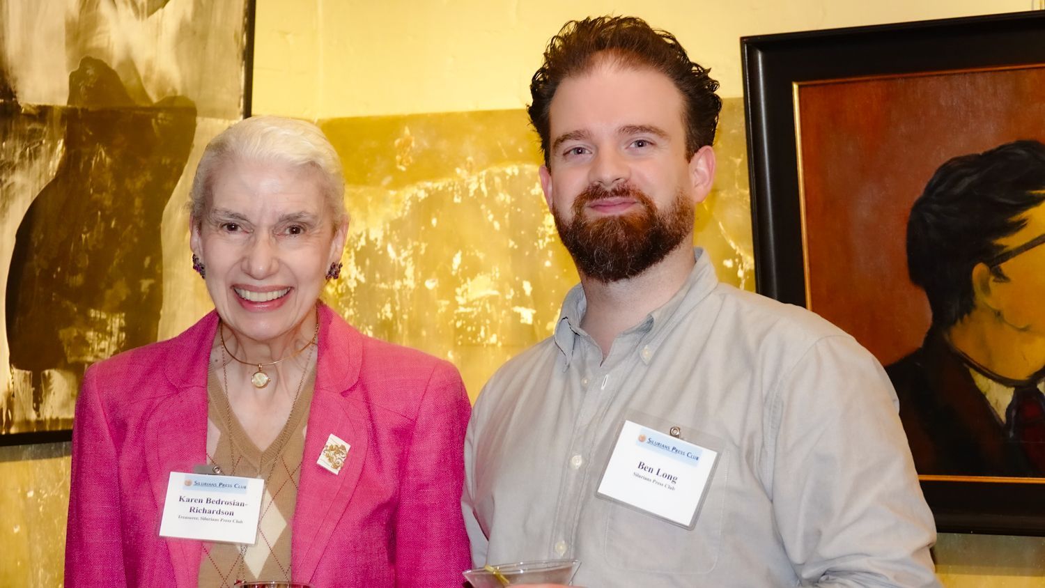 Woman in pink blazer smiles next to man with beard, both wearing name tags, posing in front of artwork.