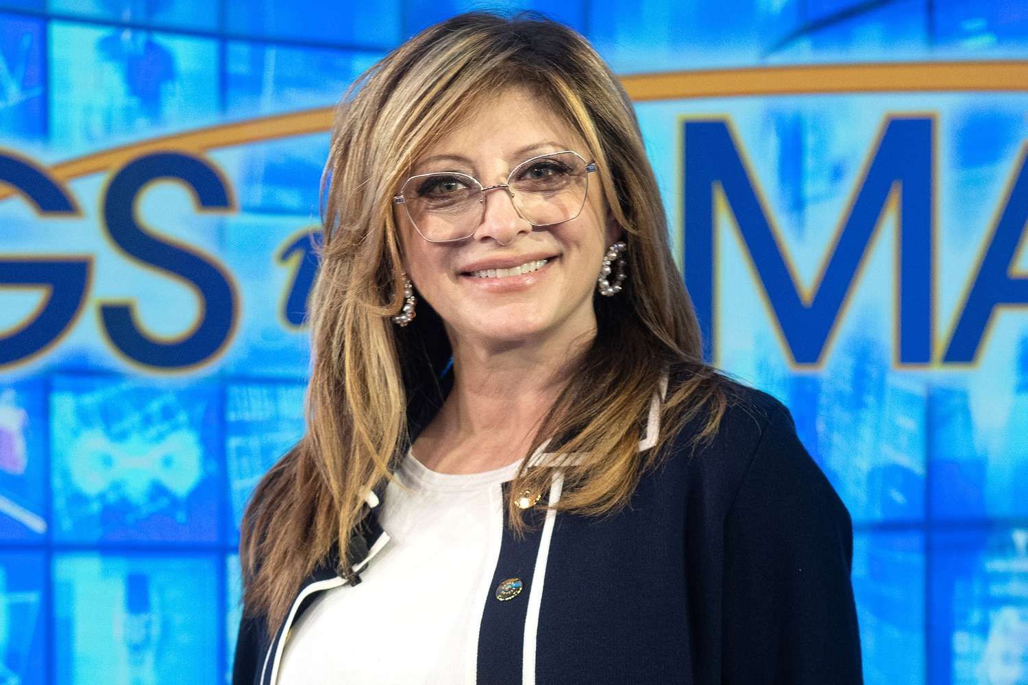 Smiling woman in glasses standing in front of a blue TV studio backdrop