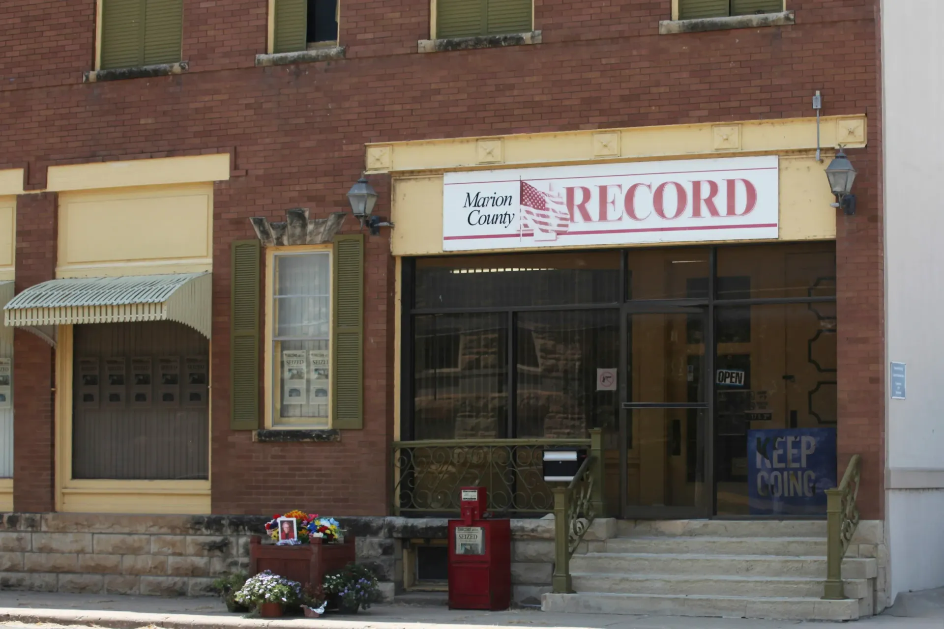 Brick storefront with a RECORD sign, steps, mailboxes, and a flower display on the sidewalk.
