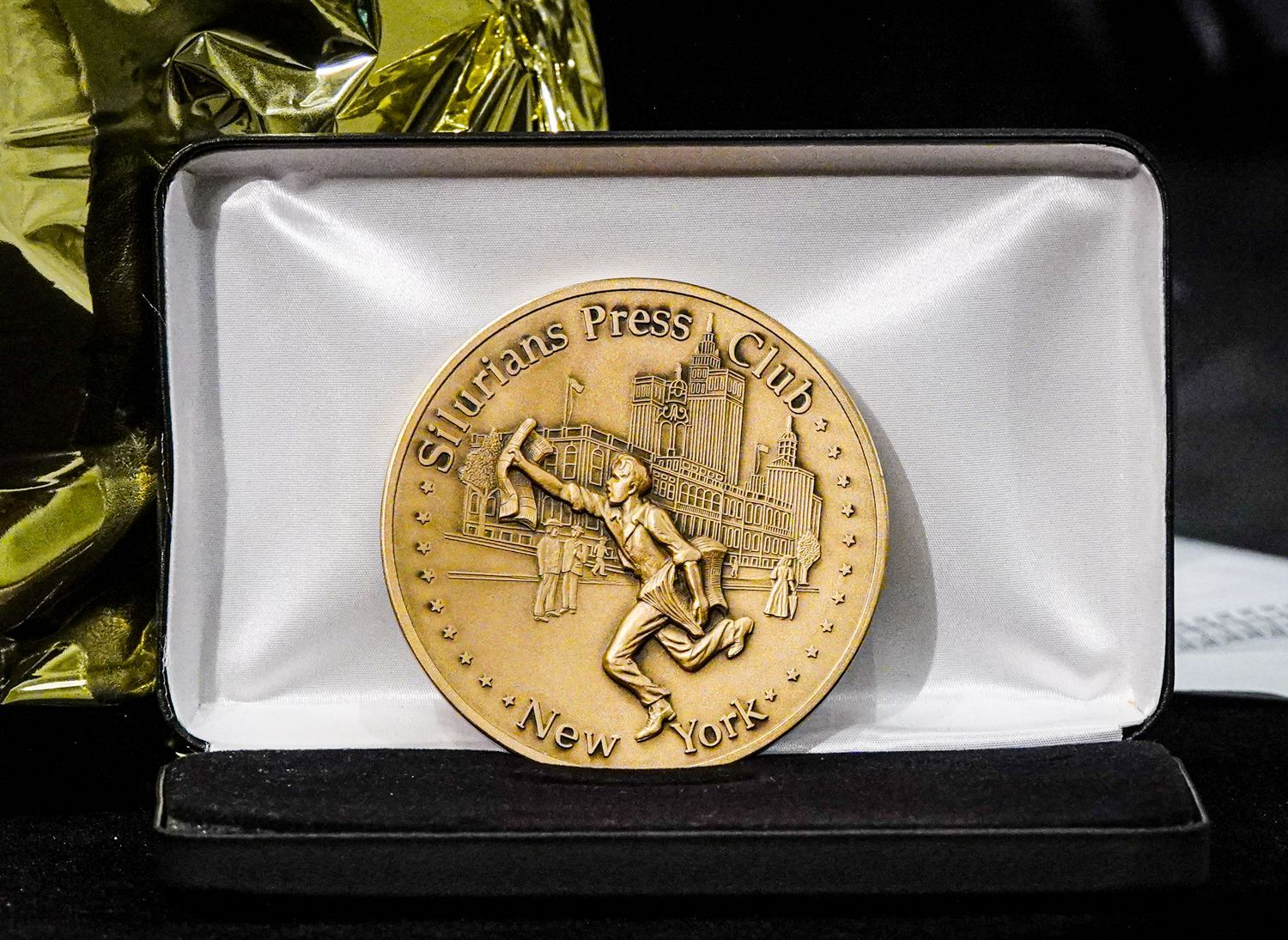 Gold medal in a black presentation box. Depicts a running figure, cityscape, and text 