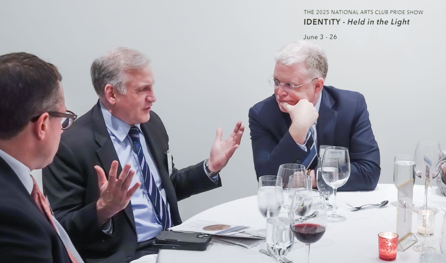 Three men in suits at a round table, conversing. One gestures with hands. Glasses and a candle are present.
