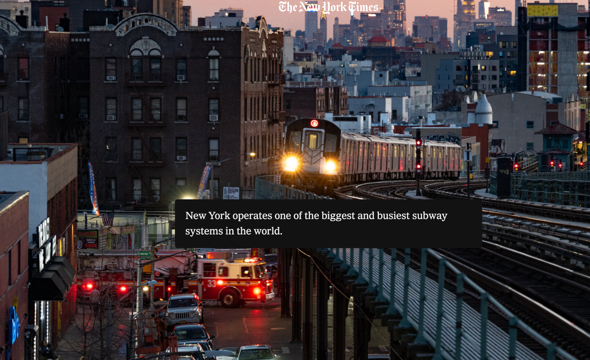 Subway train approaching elevated tracks at dusk in a city, with red lights and fire trucks below.