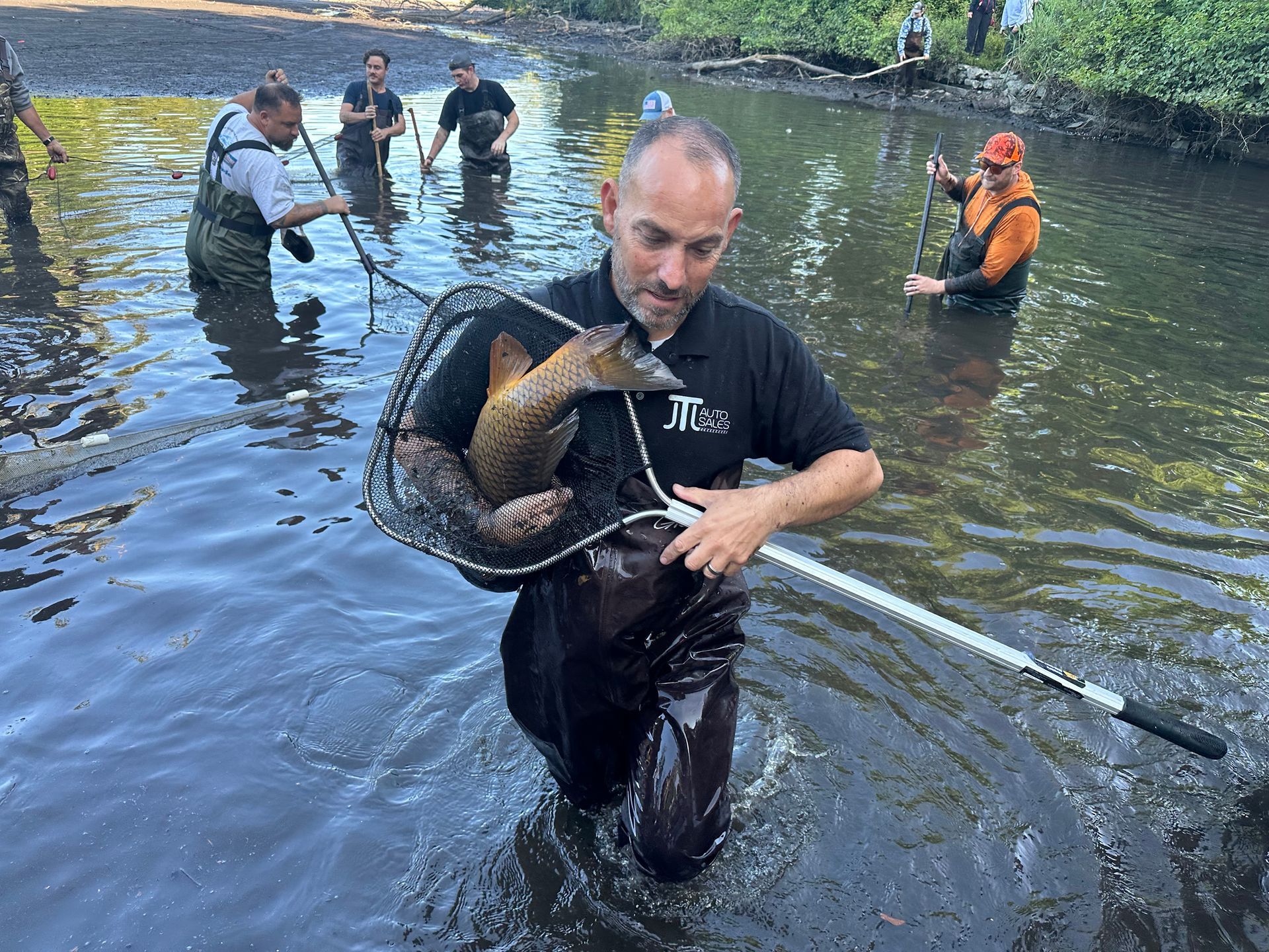 Man in waders holds a large fish in a net while in a muddy stream, other people in waders in background.