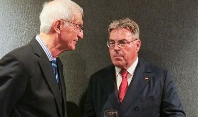Two men in suits talking; one is wearing a red tie. Dark background.