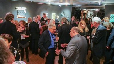 People socializing in an art gallery, some holding drinks. Dim lighting, artwork on walls.