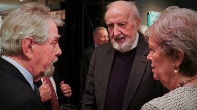 Three people conversing closely at an event. A man with a beard and a jacket is talking to a woman and another man with grey hair.