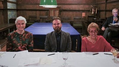 Three people seated at a table, one in the middle with a beard. They are in a wood-paneled room.