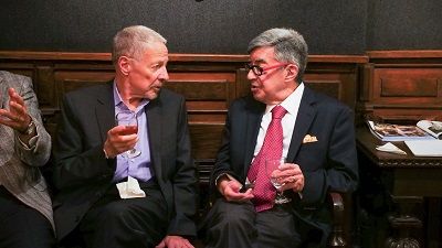 Two men seated, in conversation, holding drinks. Interior, dark wood paneling in background.