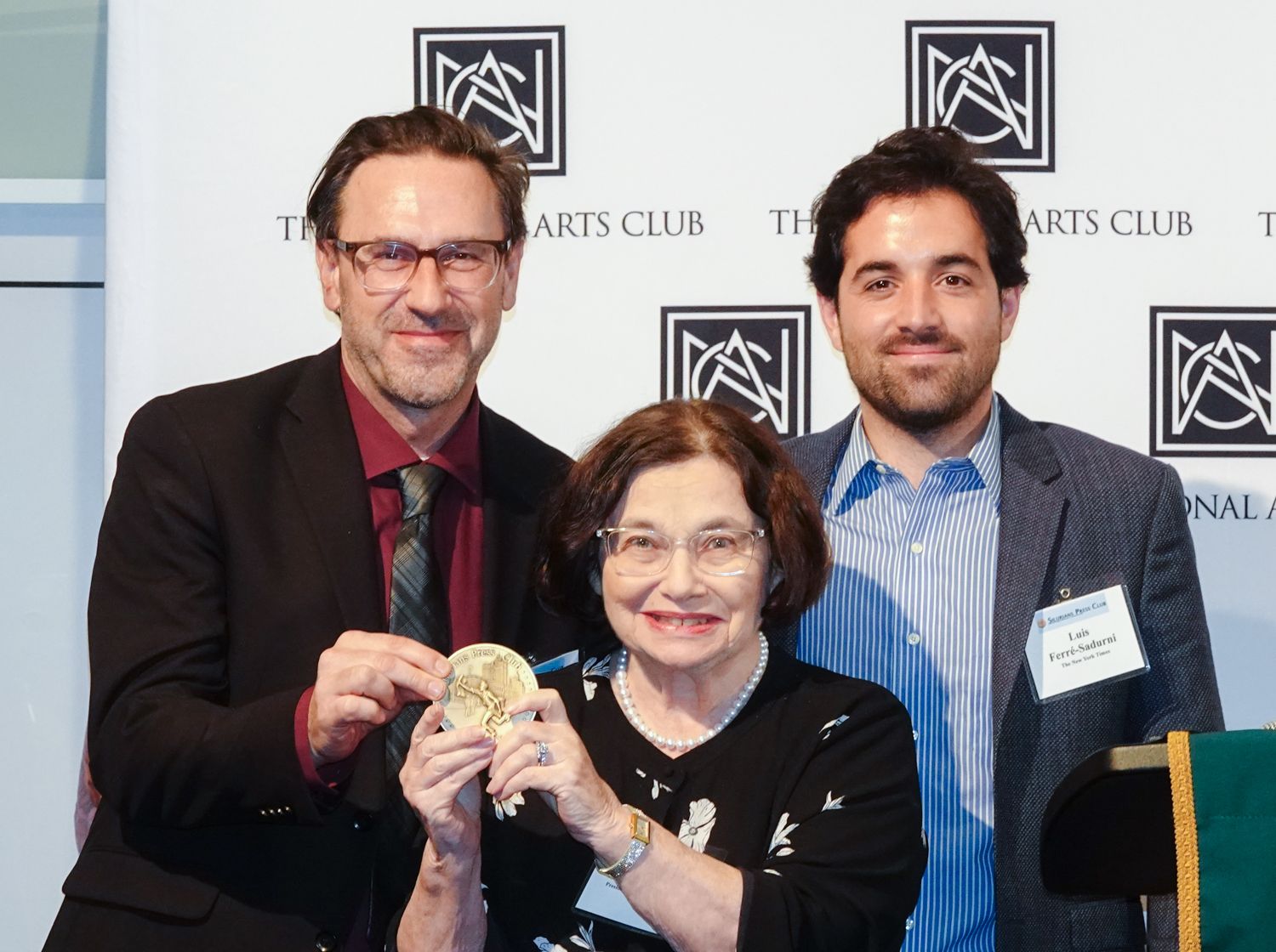 Three people at an awards ceremony; woman holds a medal, flanked by two men in front of a logo.
