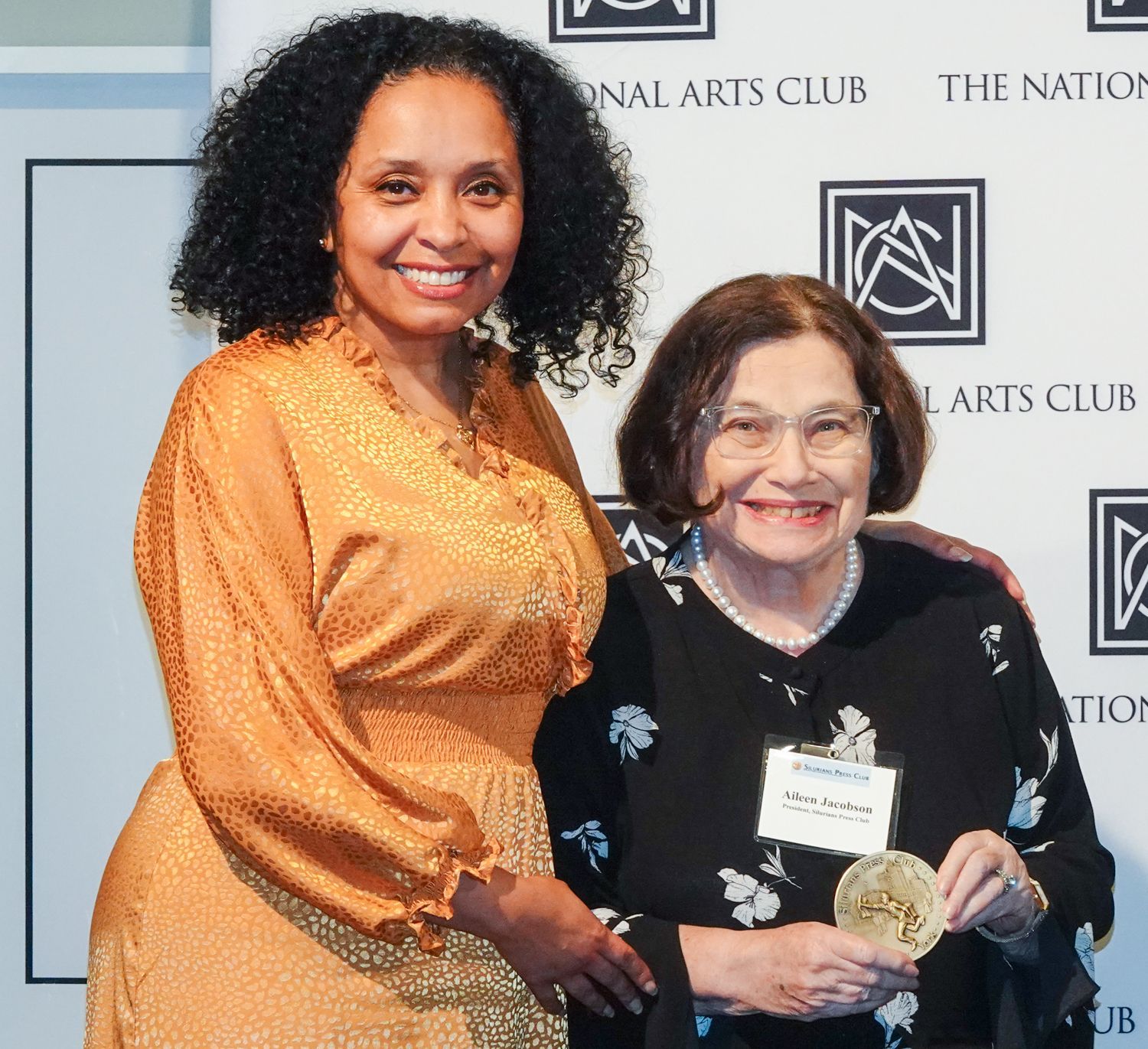 Woman in gold dress stands next to older woman holding medal at the National Arts Club.