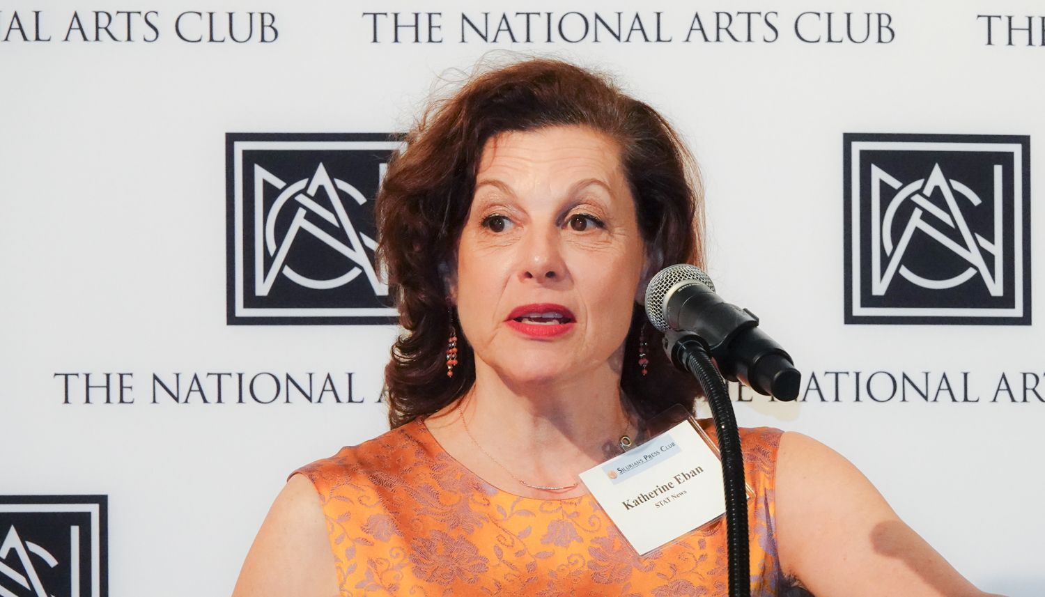 Woman speaking at a podium, at The National Arts Club. She wears an orange patterned top, and a name tag.