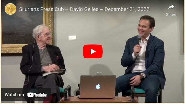 Two men in a room, one older, one younger, seated and talking; one with a microphone, laptop present.