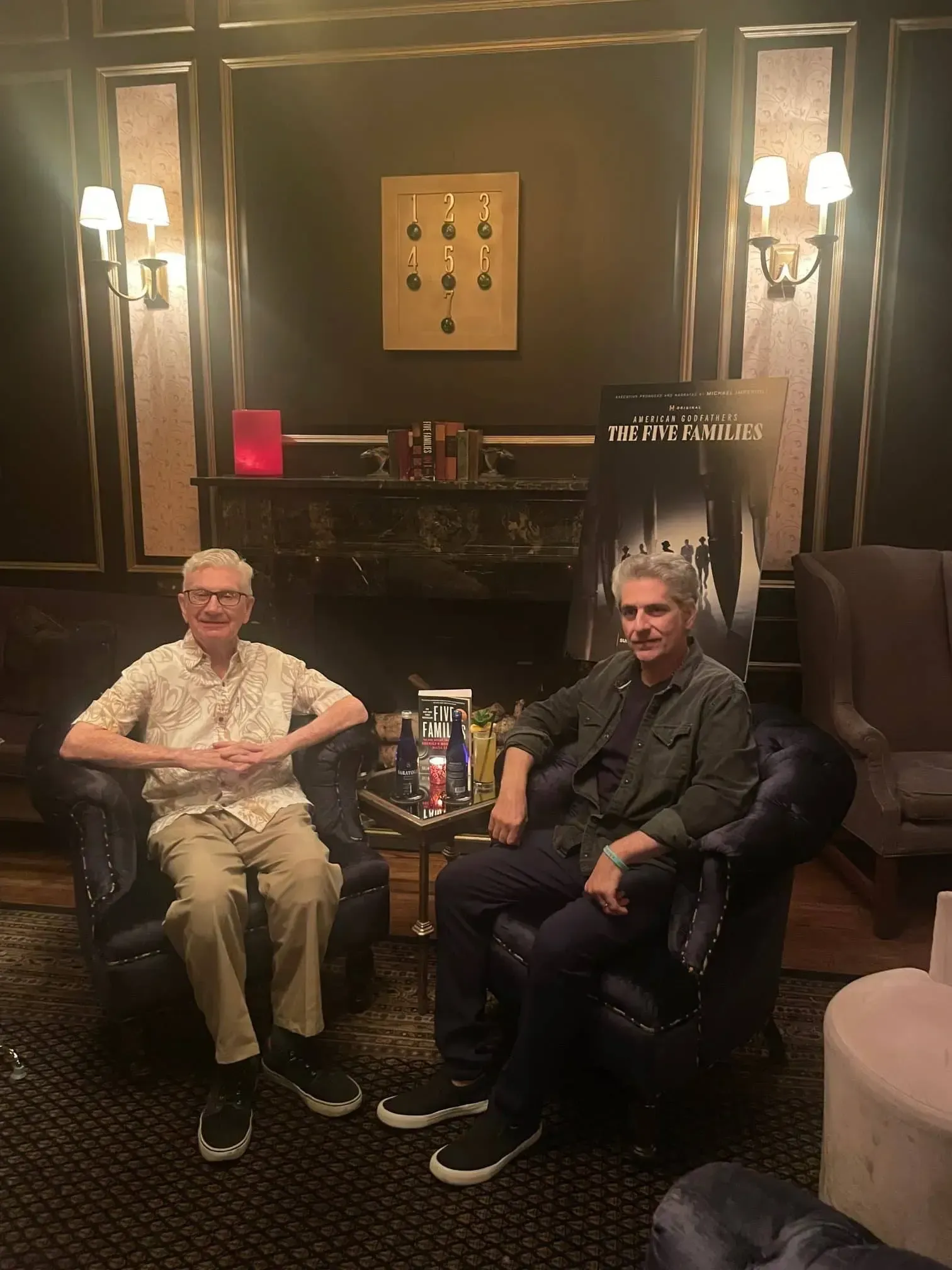 Two men sit in a dimly lit room, a movie poster in the background. They are in armchairs and talking.