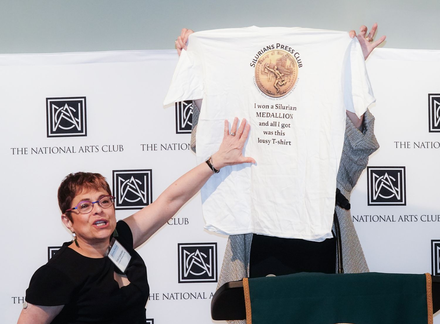 Woman holding up a white t-shirt at The National Arts Club. The shirt has a design and text.