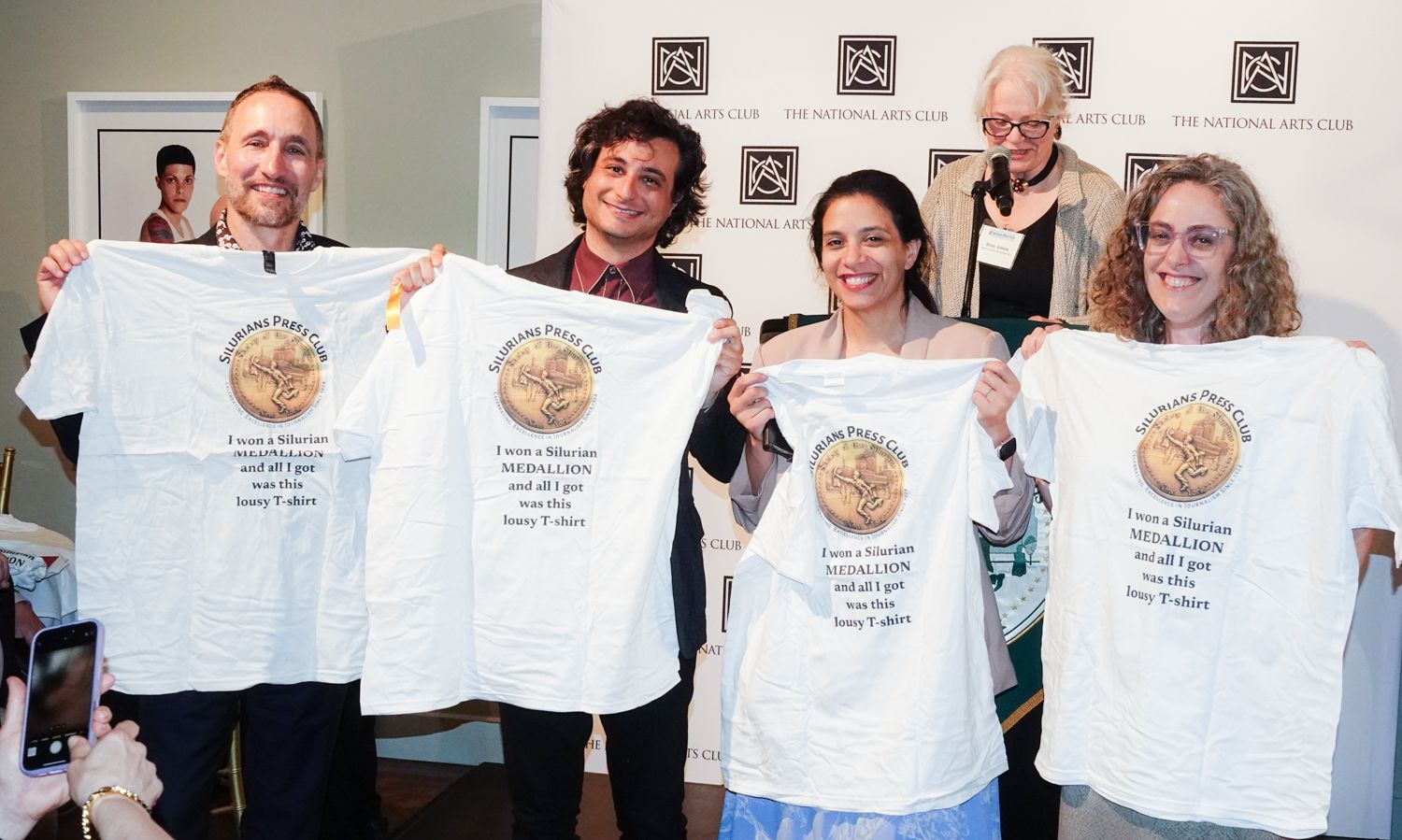 Group of people holding up white t-shirts with a graphic. They are indoors, in front of a white background.