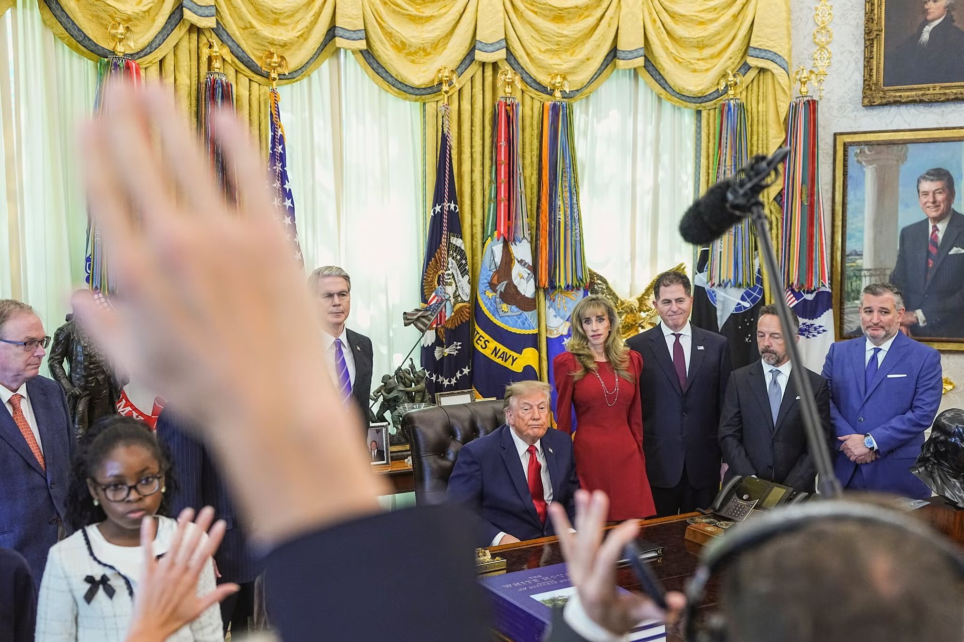Group of people in a decorated room as a crowd raises hands during a formal ceremony with flags behind them
