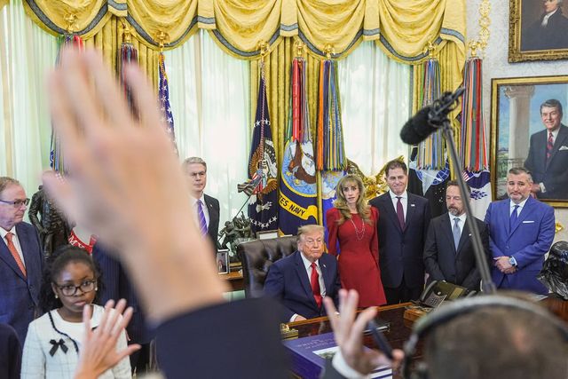 Group of people in a decorated room as a crowd raises hands during a formal ceremony with flags behind them