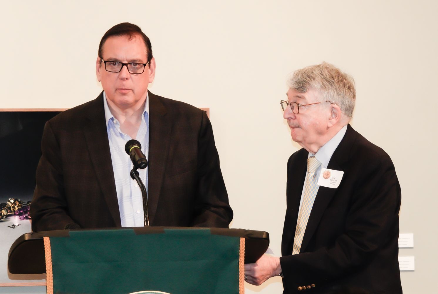 Two men at a podium. One speaks into a microphone, the other stands to the side.