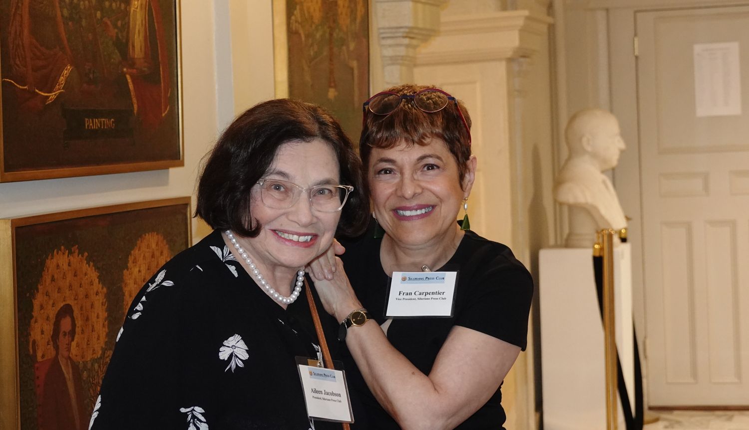 Two women smile, pose together near artwork, inside a gallery. One wears glasses, black top; the other has a name tag.