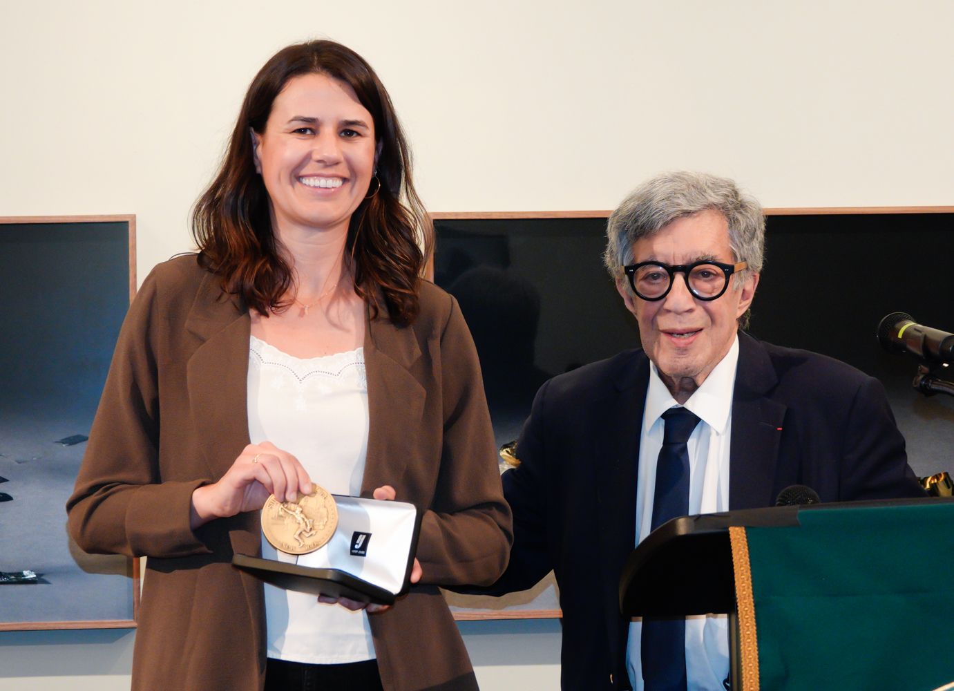 Woman in brown jacket holds award with older man beside podium. Artwork in background.