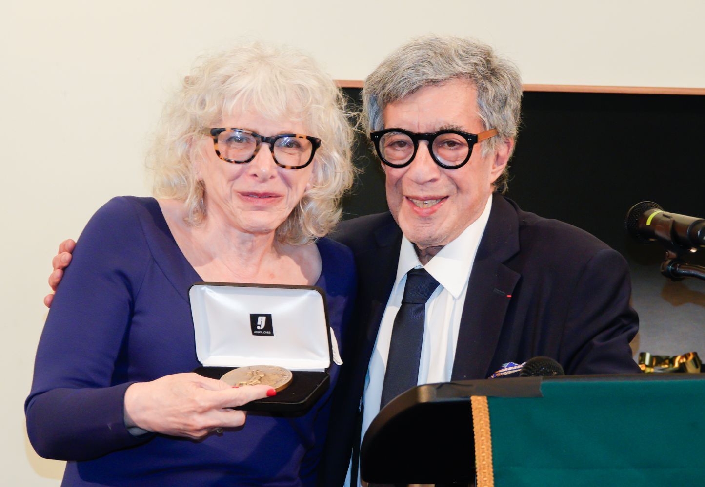 Woman holding award with man beside her at a podium. Both wear glasses.