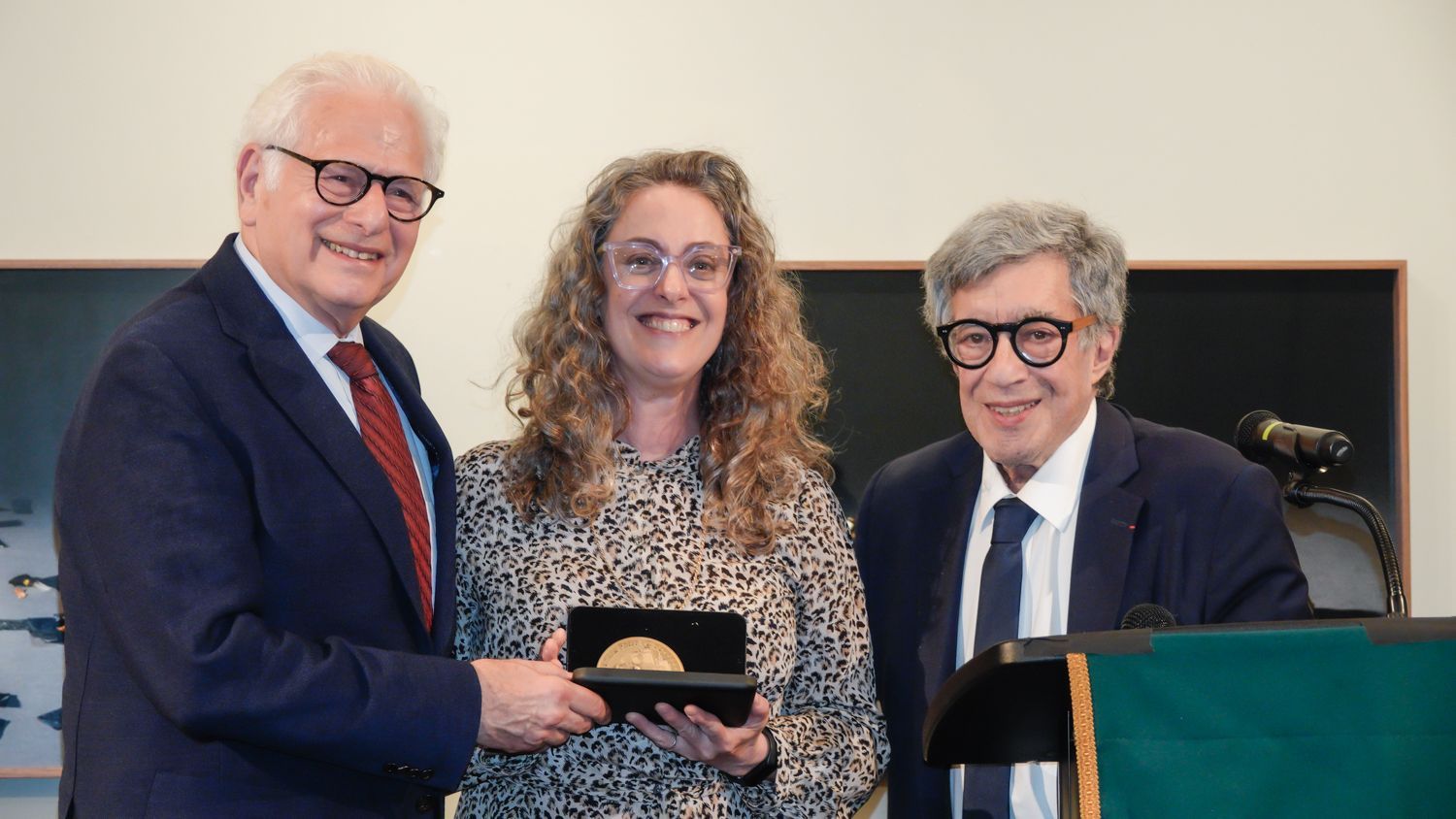 Man presenting a medal to a woman, another man stands nearby a podium; all indoors.