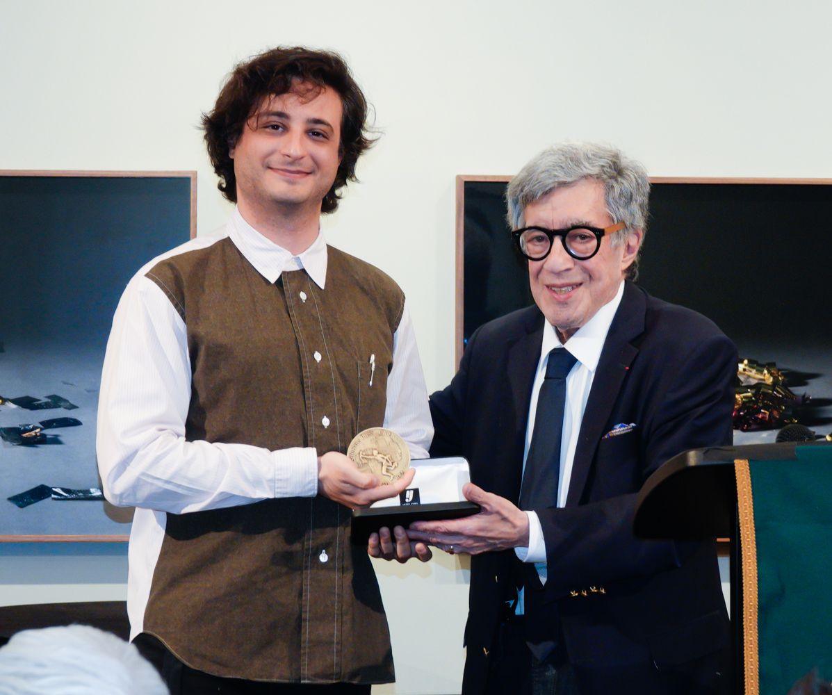 Man receiving a gold medal from another man, inside a room, smiling.
