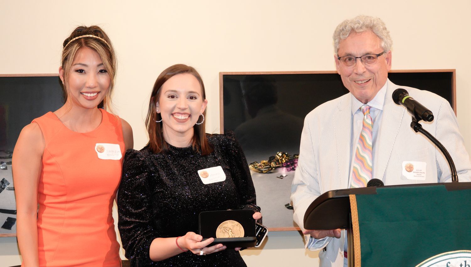 Two women and a man at a podium; one woman holds an award. They are in a well-lit room.