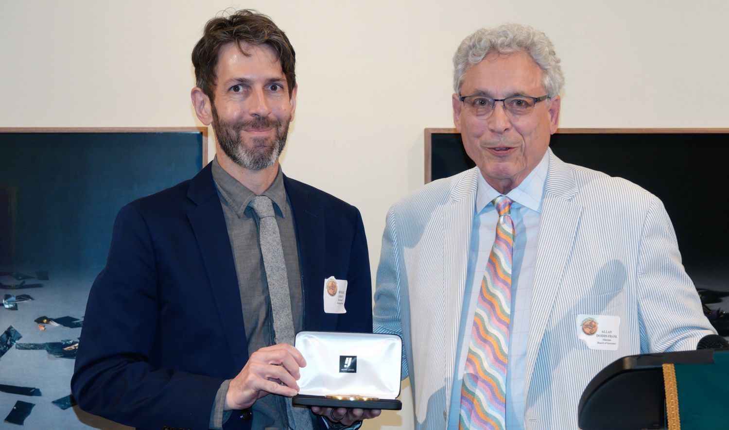 Man in a suit presents a silver box to a man in seersucker suit, both smiling. Background has artwork.