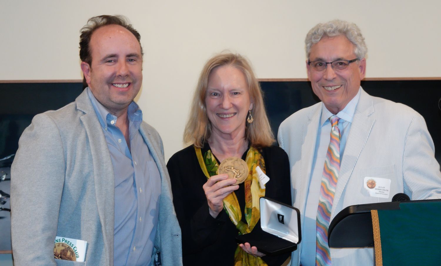 Woman holding medal with two men, smiles, at podium, light background.