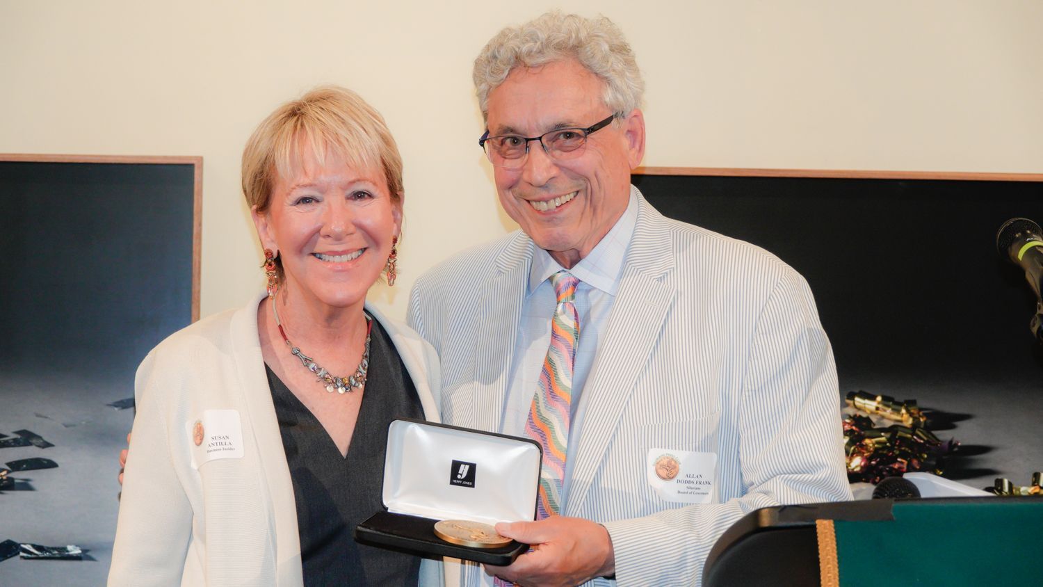 Woman presents a framed award to a man in a white striped jacket. Both smile in a well-lit room.
