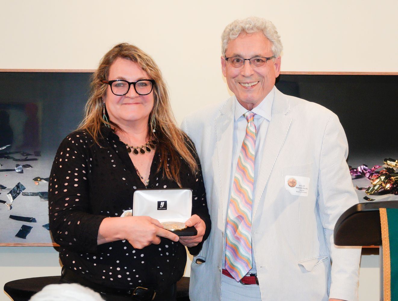 Woman holding a small gift box, smiles, with a man beside her in a striped blazer.