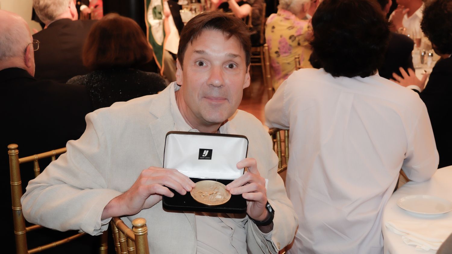 Man in cream blazer holding open box with a gold medal at an event.