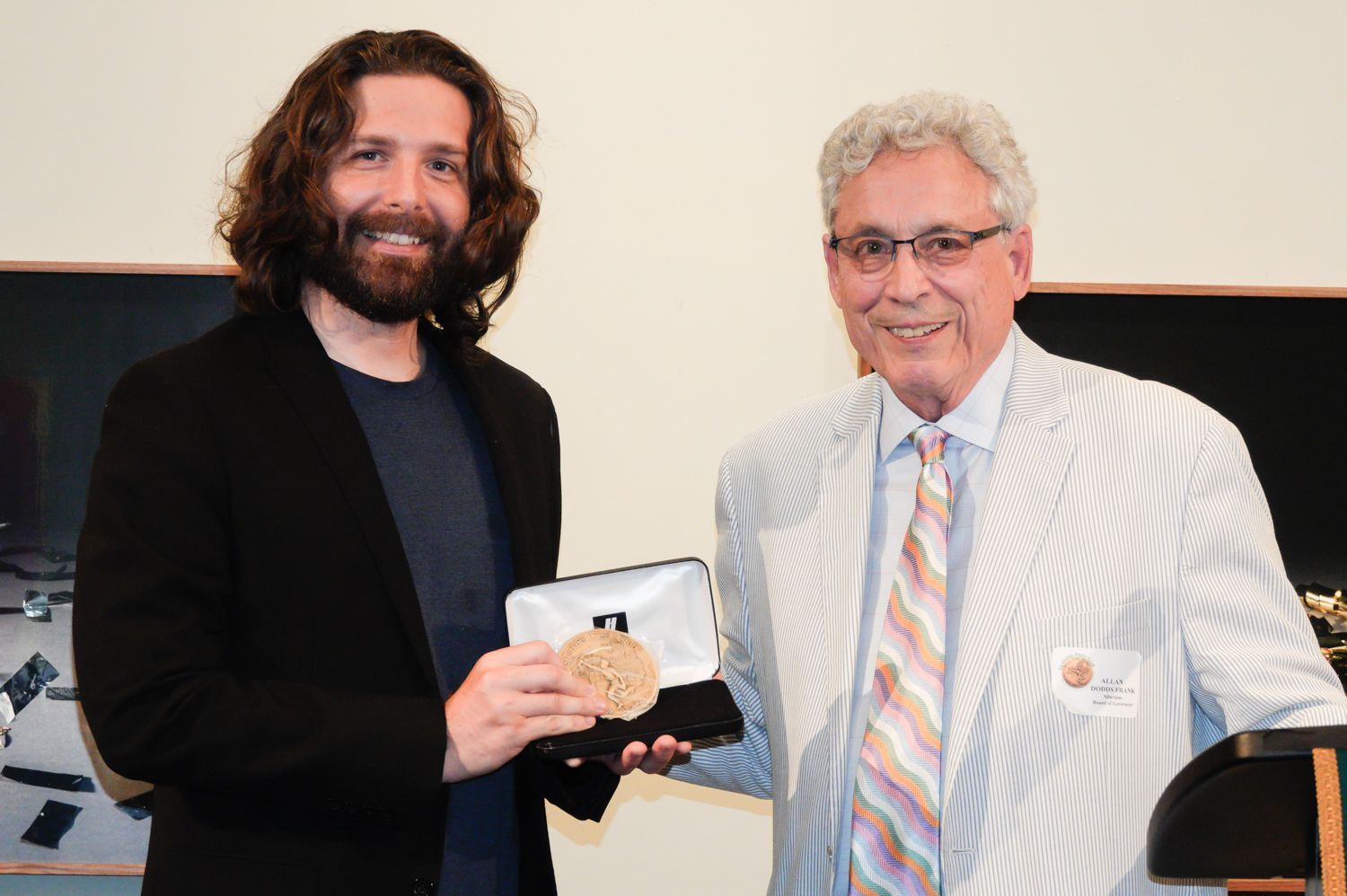 Man with long hair holds a medal, presented by an older man in a striped jacket, at a podium.