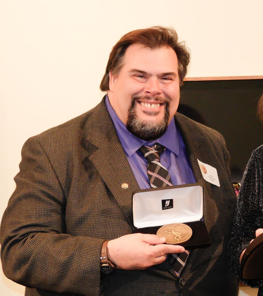 Man smiling, holding an open box with a medal, wearing a suit and tie.