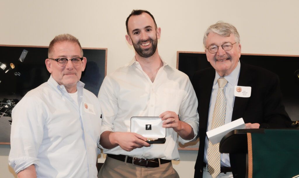 Three men at a podium; one holding a small box. They are smiling and appear to be at an awards ceremony.