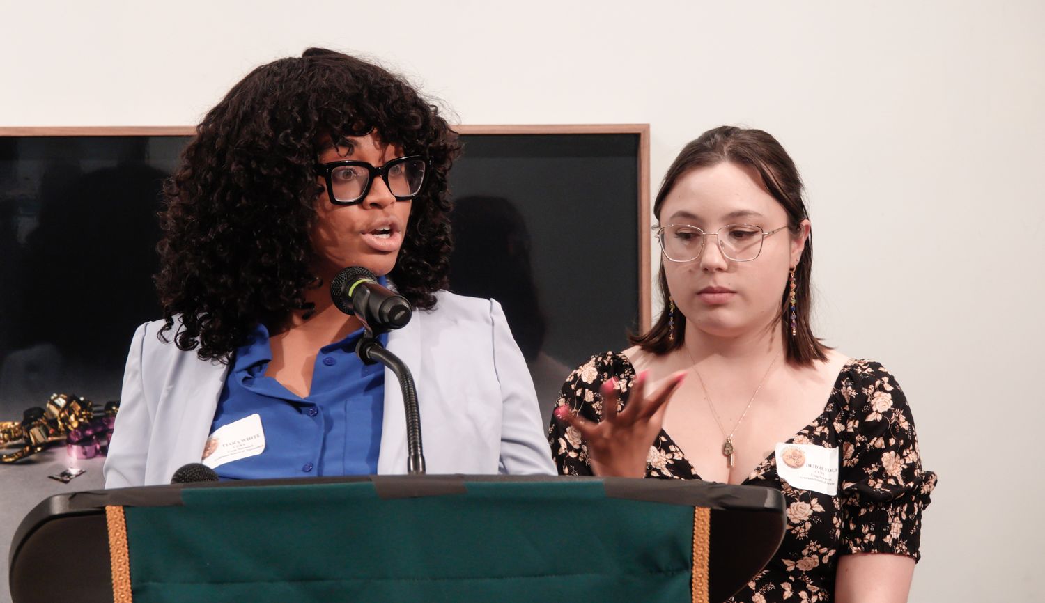 Two women at a podium, one speaking into a microphone, the other gesturing; indoor setting.