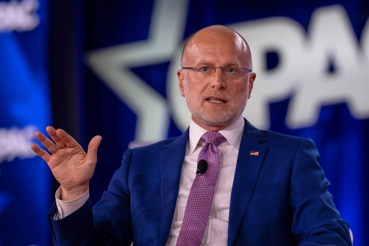Man in blue suit speaking at a CPAC stage, gesturing with one hand and wearing a pink tie.