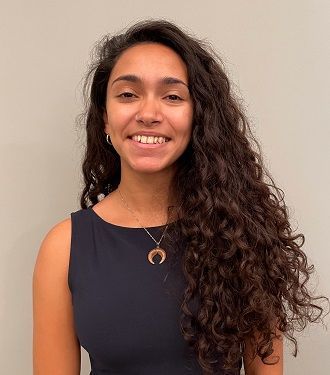 Woman with curly dark hair, smiling, wearing a dark sleeveless top and necklace, against a gray wall.